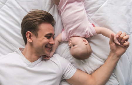 Top view of handsome young dad and his cute baby looking at each other while lying on bed