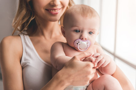Beautiful young mom is holding her cute baby in arms and smiling while standing near the windowの写真素材