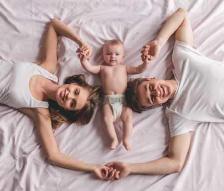 Top view of beautiful young parents and their cute baby holding hands, looking at camera and smiling...