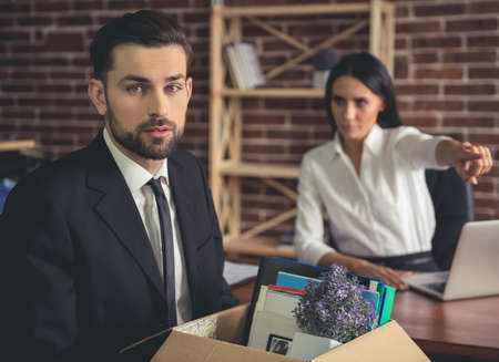 Getting fired. Handsome businessman in suit is holding a box with his stuff, woman in the background is pointing awayの写真素材