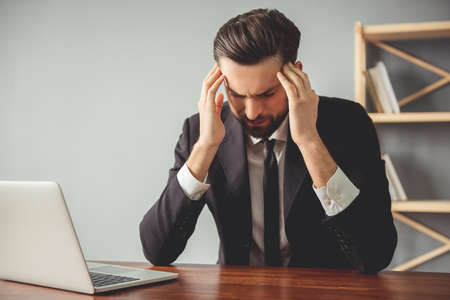 Handsome tired businessman in suit is touching his temples while working in his officeの写真素材