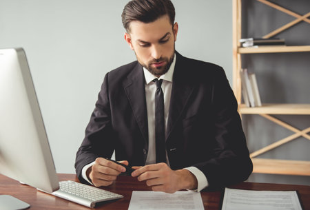 Handsome young businessman in suit is examining documents and using a computer while working in his...