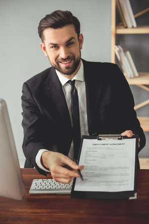 Handsome young businessman in suit is offering to sign an employment agreement and smiling while sitting at the table in his officeの写真素材