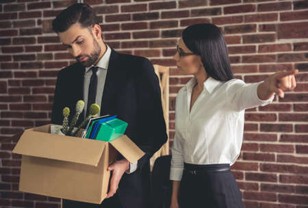 Beautiful female boss is firing young businessman and pointing away while he is holding a box with his stuffの写真素材