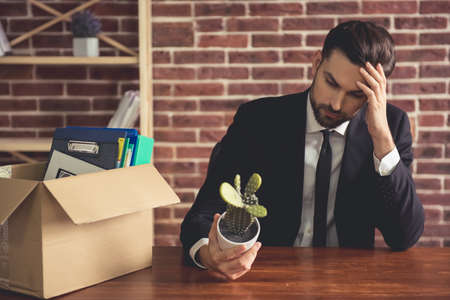 Getting fired. Handsome businessman in suit is sitting sadly at the table in office near the box with his stuffの写真素材