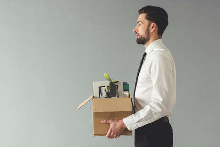 Getting fired. Side view of handsome businessman in formal wear holding a box with his stuff, on gray backgroundの写真素材