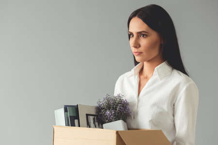 Getting fired. Beautiful young business woman is holding a box with her stuff and looking sadly, on gray backgroundの写真素材