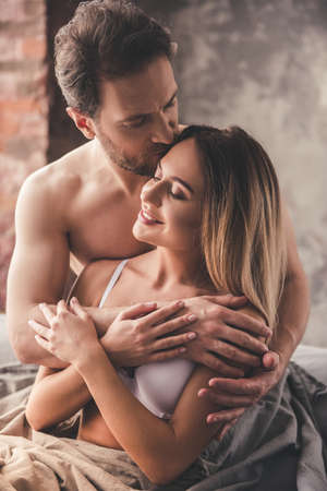 Beautiful couple is smiling and hugging while spending time together in bed at home. Man is kissing his girlfriendの写真素材