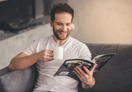 Handsome man is reading a magazine, holding a cup and smiling while resting at homeの写真素材
