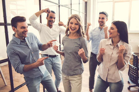 Successful business team is raising hands in fists and smiling while standing in officeの写真素材