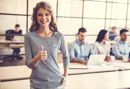 Beautiful business lady is showing Ok sign and smiling, in the background her colleagues are working in officeの写真素材