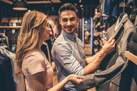 Beautiful couple is choosing formal suit and smiling while doing shopping in the male store