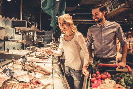 Beautiful young couple is choosing fish and smiling while doing shopping at the supermarketの写真素材