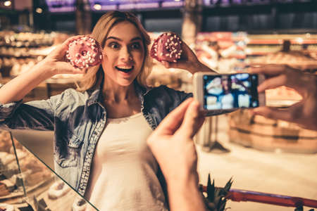 Couple at the supermarket. Girl is holding donuts and posing while her boyfriend is taking a photoの写真素材