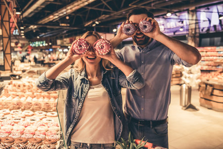 Beautiful couple is holding donuts and smiling while doing shopping at the supermarketの写真素材