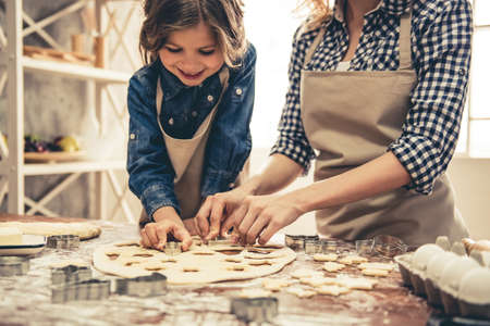 Cute little girl and her beautiful mom in aprons are smiling while preparing cookies using cookie cutters in the kitchenの写真素材