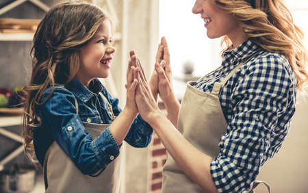 Cute little girl and her beautiful mom in aprons and chef hats are holding hands and smiling while baking in the kitchenの写真素材