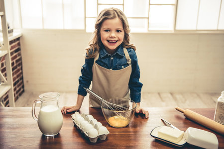 Cute little girl in apron is looking at camera and smiling while whisking eggs for baking in the kitchenの写真素材