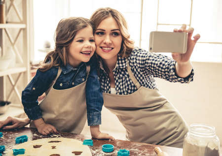 Cute little girl and her beautiful mom in aprons are doing selfie and smiling while preparing cookies using cookie cutters in the kitchenの写真素材