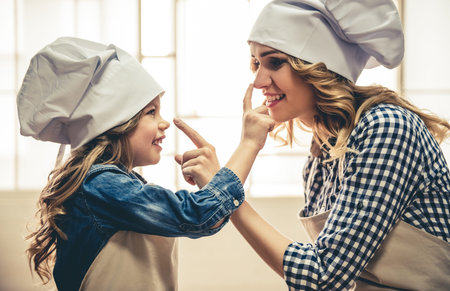 Cute little girl and her beautiful mom in aprons and chef hats are touching noses and smiling while kneading the dough in the kitchenの写真素材