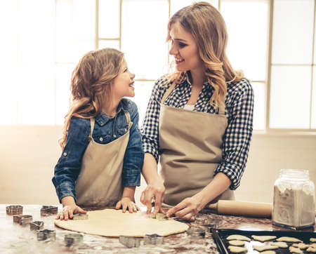 Cute little girl and her beautiful mom in aprons are smiling while preparing cookies using cookie cutters in the kitchenの写真素材