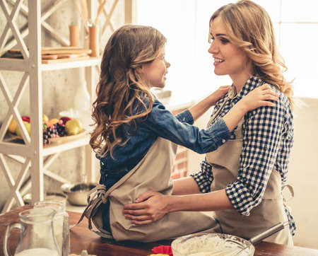 Cute little girl and her beautiful young mom are hugging, looking each other and smiling while baking in kitchenの写真素材