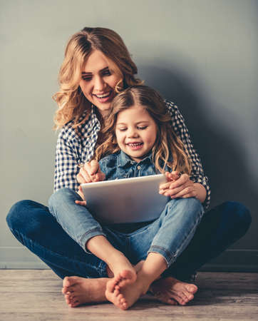 Cute little girl and her beautiful young mom are sitting together on the floor, using a digital tablet and smiling, on gray backgroundの写真素材