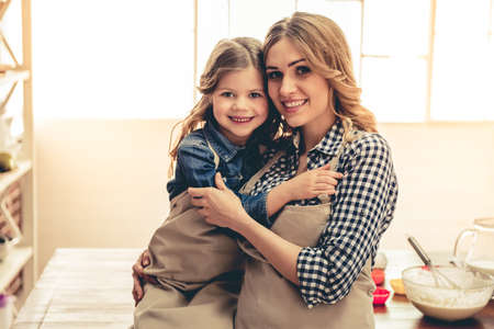 Cute little girl and her beautiful young mom are hugging, looking at camera and smiling while baking in kitchenの写真素材