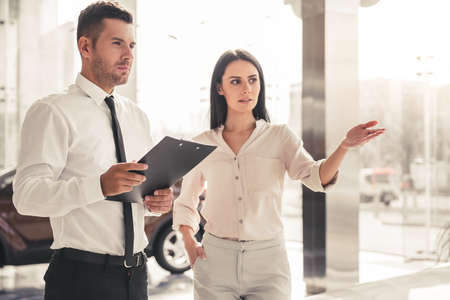 Beautiful young woman is talking to handsome car dealership worker while choosing a car in dealershipの写真素材