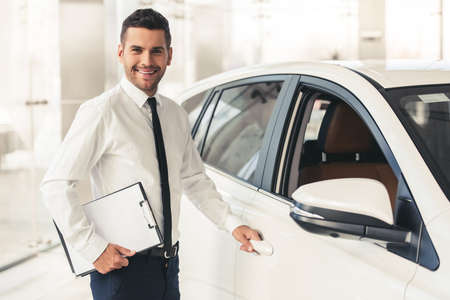 Handsome car dealership worker in suit is holding a folder, looking at camera and smiling while standing near the carの写真素材