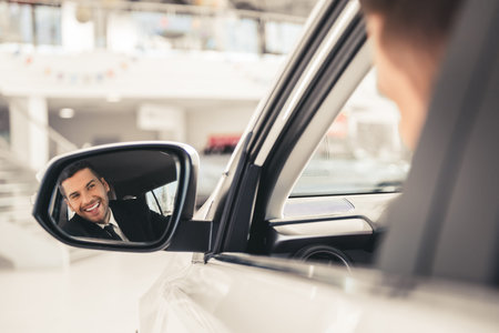 Handsome businessman in suit is looking into the car side mirror and smiling while sitting in a new...