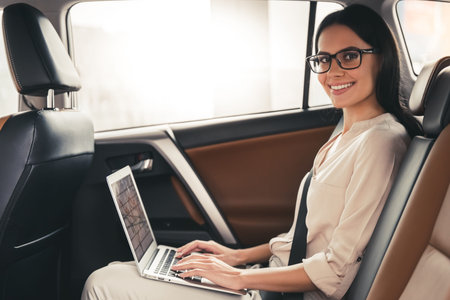 Beautiful business woman in eyeglasses is using a laptop and smiling while sitting on back seat in the carの写真素材