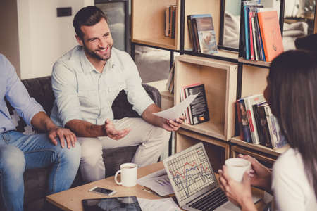 Successful young business people are using gadgets, sharing ideas, drinking coffee and smiling while working in officeの写真素材