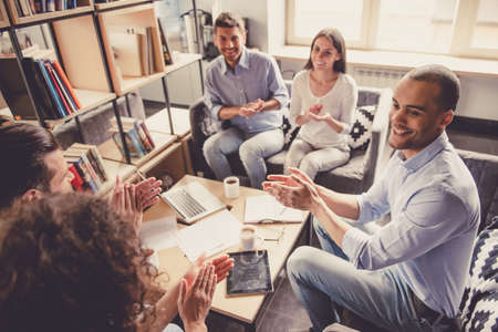 Successful young business people are using gadgets, sharing ideas, applauding and smiling while working in officeの写真素材