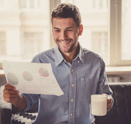 Handsome businessman is drinking coffee, studying document and smiling while working in officeの写真素材