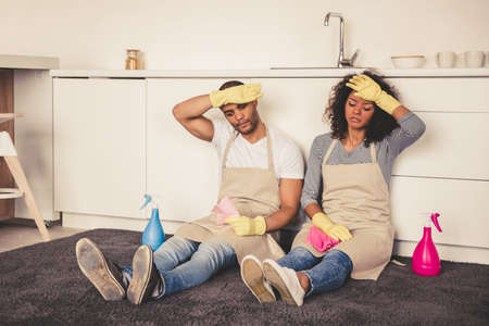 Beautiful tired young Afro American couple is wiping foreheads while sitting on the floor in kitchen after cleaning itの写真素材
