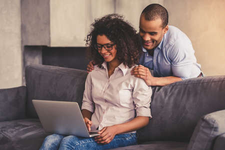 Beautiful young Afro American couple on couch at home. Business woman is using a laptop while her boyfriend is doing her massageの写真素材