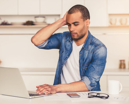 Handsome tired Afro American businessman is working with a laptop in kitchen