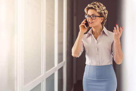 Beautiful psychotherapist in suit and eyeglasses is talking on the mobile phone and smiling while standing in officeの写真素材