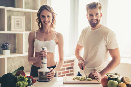 Beautiful young couple is looking at camera and smiling while cooking healthy food in kitchen at homeの写真素材