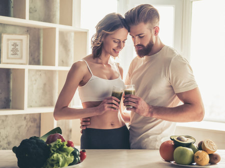 Beautiful young couple is hugging and smiling while drinking nutritious cocktails in kitchen at homeの写真素材