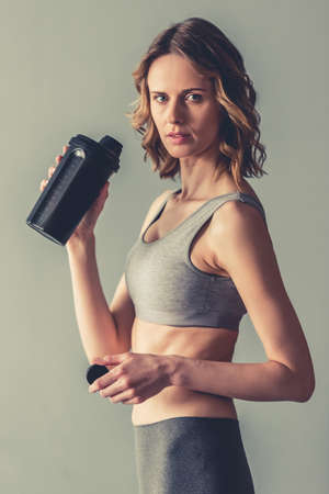 Beautiful young sportswoman is holding a shaker with sport nutrition and looking at camera, on gray backgroundの写真素材