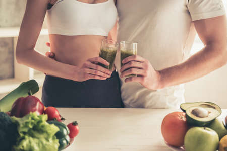 Beautiful young couple is hugging and smiling while drinking nutritious cocktails in kitchen at homeの写真素材