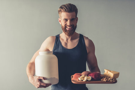 Handsome young bearded sportsman is holding a bottle with sport nutrition and a wooden tray with...