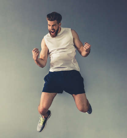 Handsome young bearded sportsman is showing muscles, jumping and smiling, on gray backgroundの写真素材