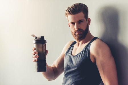 Handsome young bearded sportsman is holding bottle of water and looking at camera, on gray...