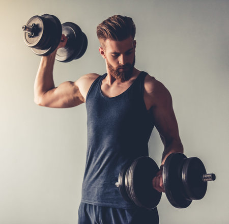 Handsome young bearded sportsman is working out with dumbbells, on gray backgroundの写真素材