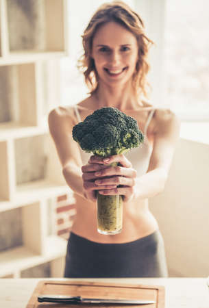 Beautiful young sportswoman is looking at camera and smiling while cooking healthy food in kitchen at homeの写真素材