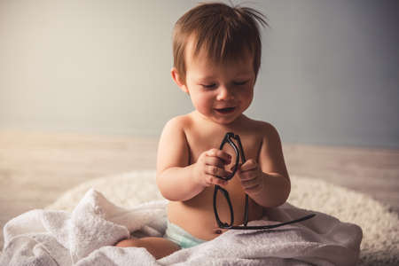 Cute baby boy in diaper is playing with eyeglasses and smiling while sitting on the carpet at homeの写真素材