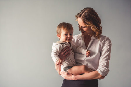 Beautiful business mom in suit is playing with her cute baby boy and smiling, on gray backgroundの写真素材